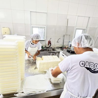 A imagem mostra dois trabalhadores em uma fábrica de queijos, imersos no processo de produção.(1)