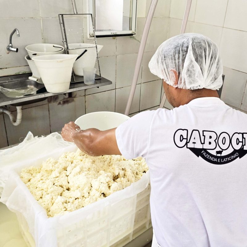 Um profissional de cozinha, vestido com uniforme branco, manipula coalhada em um grande recipiente. O ambiente é uma fábrica de alimentos, com equipamentos de aço inoxidável.